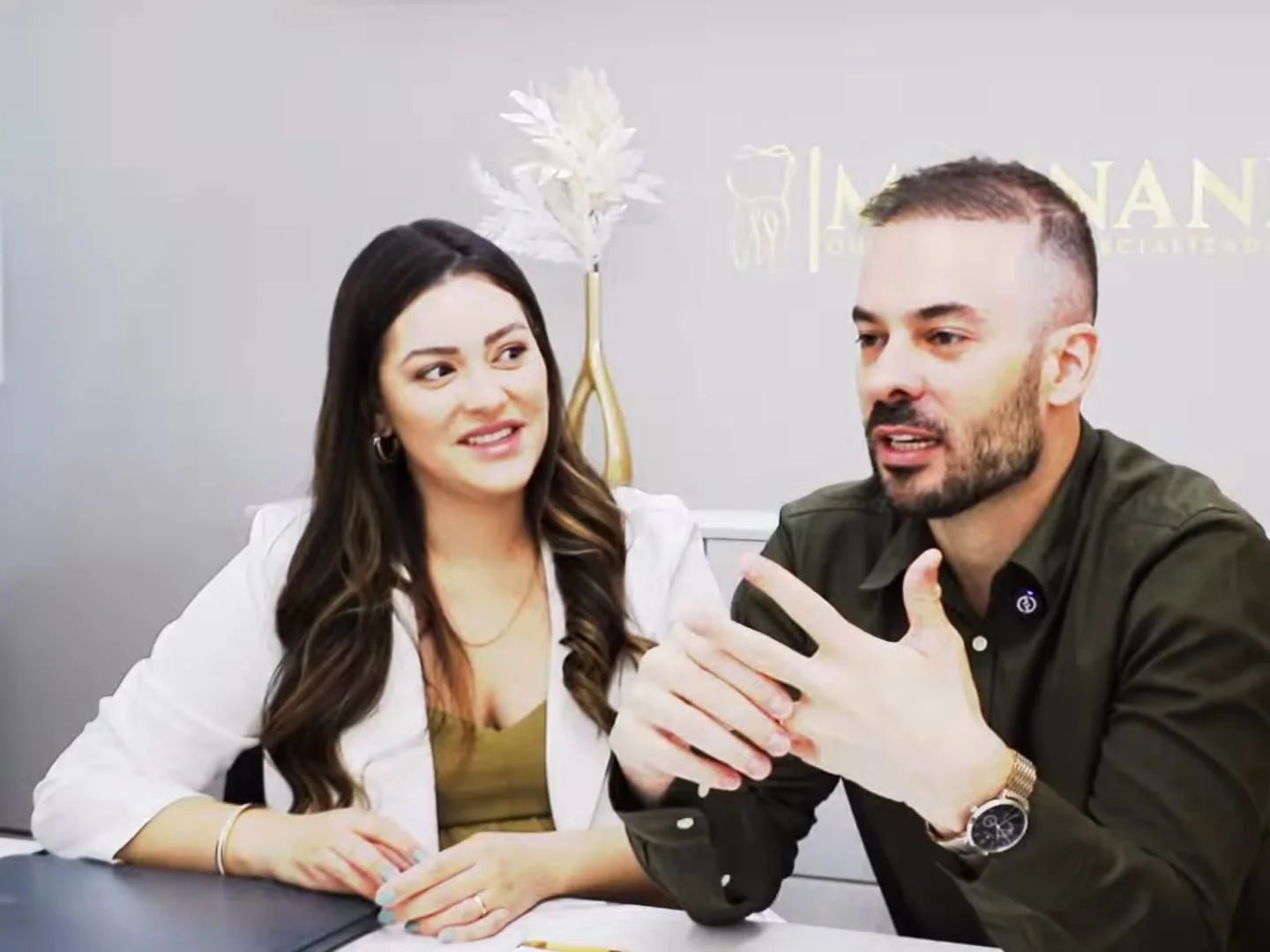 Dr. Guilherme Magnani explica com as mãos, sentado à mesa do consultório; ao lado, a Dra. Yngrid Collete olha para ele e sorri