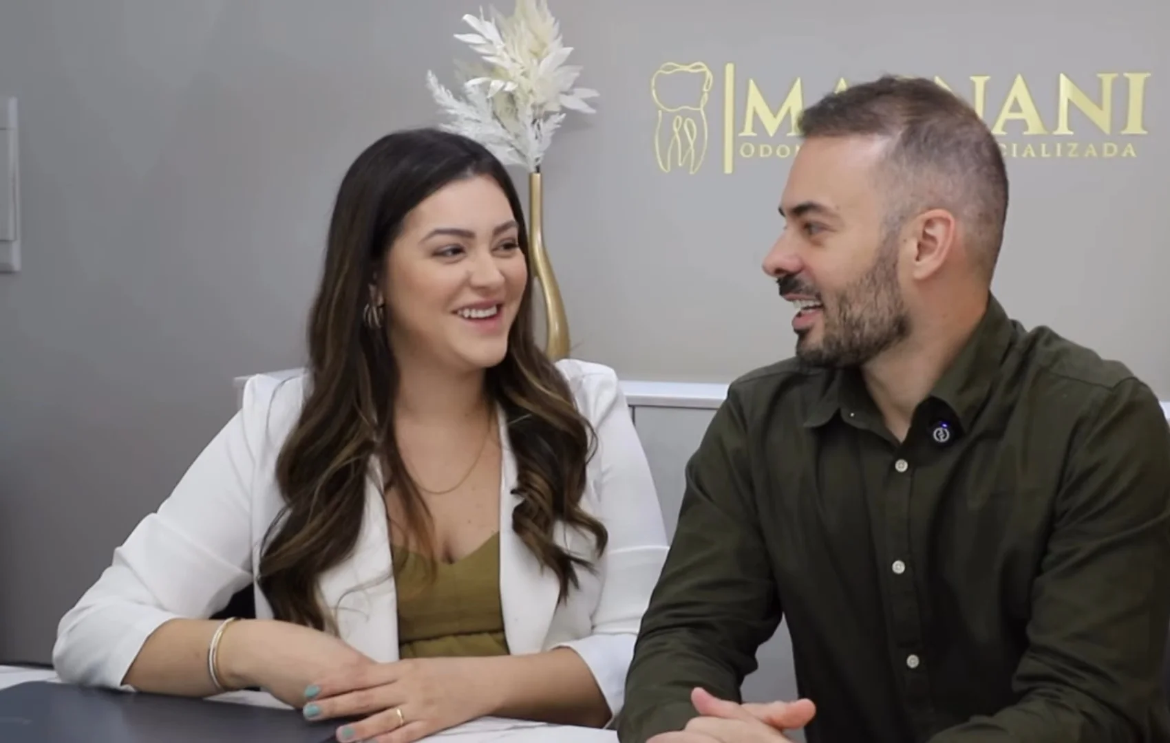 Dra. Yngrid Collete e Dr. Guilherme Magnani rindo, sentados à mesa do consultório; na parede atrás deles, o logo dourado da Magnani Odontologia Especializada