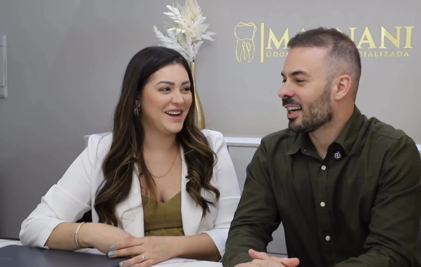 Dra. Yngrid Collete e Dr. Guilherme Magnani rindo, sentados lado a lado à mesa do consultório. Na parede atrás deles, o logo dourado da clínica.