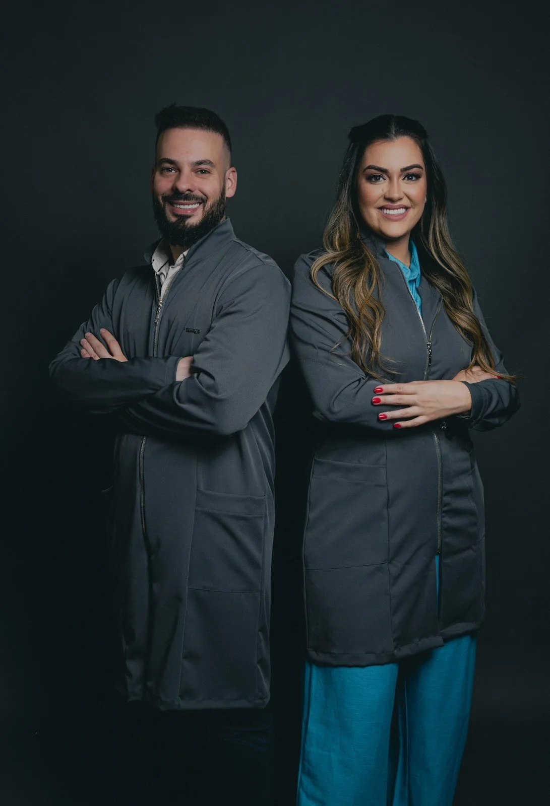 Dr. Guilherme Magnani e Dra. Yngrid Collete de costas um para o outro, de braços cruzados e sorrindo, de jaleco cinza